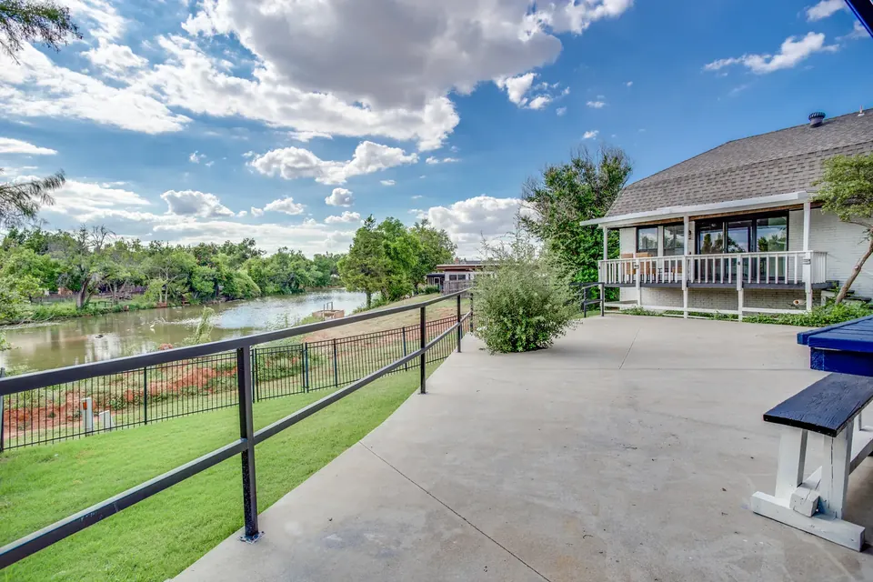 Spacious back patio with black metal railing overlooking a tree-lined pond, with the white brick home's covered deck under a blue sky at Brompton Manor, Oklahoma City