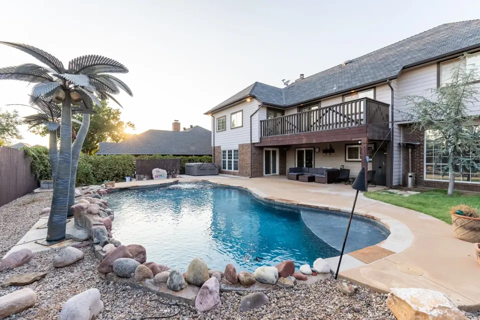Backyard freeform pool with rock border and metal palm sculptures beside a two-story home with balcony deck at sunset at Gaillardia Manor, Oklahoma City
