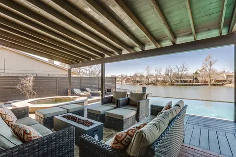 Covered patio lounge with wicker sofas and a fire pit table overlooking the infinity-edge pool and lake at Lakefront Manor, Oklahoma City