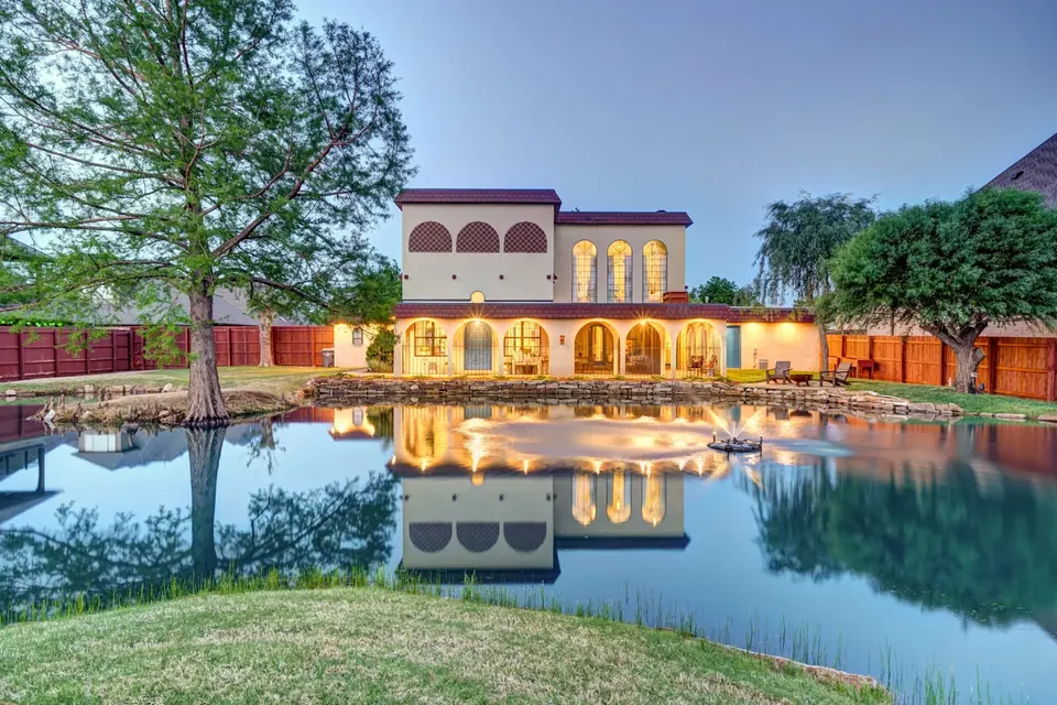 Twilight view of the Spanish-style home with lit arched loggia and fountain reflected in the backyard pond at Lakeside Manor, Oklahoma City