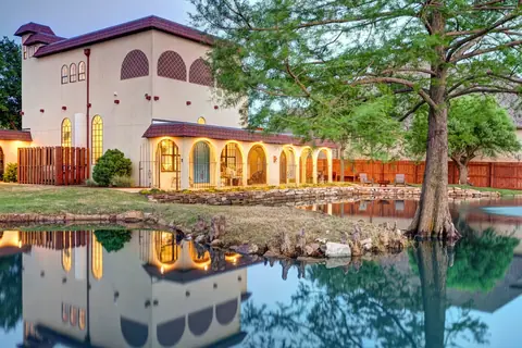 Dusk view of the lamp-lit arched veranda and bald cypress tree mirrored in the glassy backyard pond at Lakeside Manor, Oklahoma City