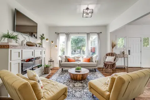 Bright living room with two mustard-yellow lounge chairs, beige sofa with orange accent pillows, electric fireplace, and wall-mounted TV at Plaza Manor, Oklahoma City