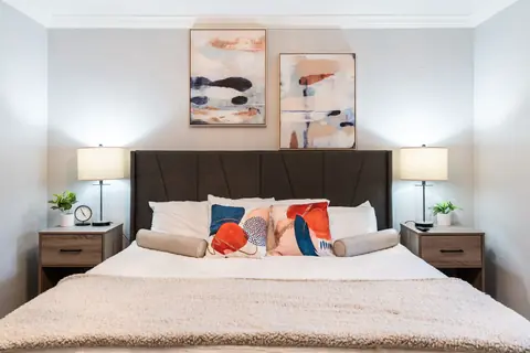 King bed styled with bolster and accent pillows beneath two abstract art prints and matching nightstand lamps at Quail Creek Manor, Oklahoma City