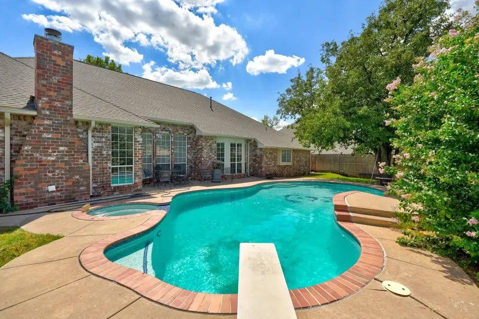 Freeform backyard pool with diving board and attached round spa beside the brick house, under blue sky with flowering crepe myrtle at Remington Manor, Oklahoma City