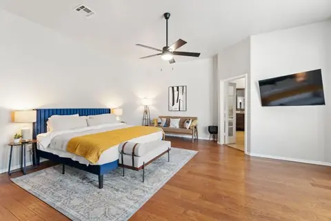 Spacious bedroom with a navy upholstered bed, yellow throw blanket, bench, loveseat, wall-mounted TV, and wood floors at Remington Manor, Oklahoma City