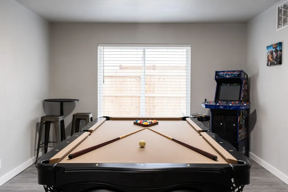 Game room with a black pool table, racked billiard balls, NFL Blitz arcade cabinet, and pub table with metal stools at The Village Manor, Oklahoma City
