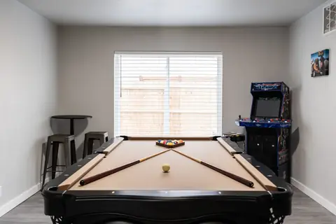Game room with a black pool table, racked billiard balls, NFL Blitz arcade cabinet, and pub table with metal stools at The Village Manor, Oklahoma City