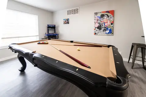 Angled view of the game room's black pool table with two cues and racked balls, arcade cabinet and abstract art on the wall at The Village Manor, Oklahoma City