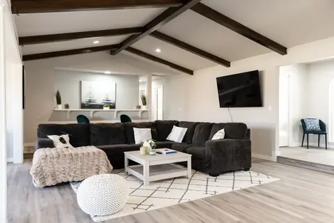 Living room with charcoal sectional, wall-mounted TV, knit pouf, and vaulted ceiling with dark wood beams at Will Rogers Manor, Oklahoma City