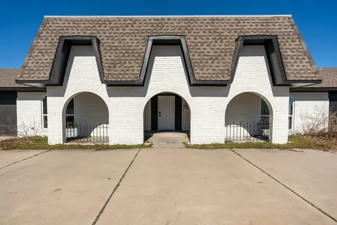 White-painted brick front exterior with three dramatic arched porch openings, mansard-style shingle roof, and wide concrete driveway under a blue sky at Will Rogers Manor, Oklahoma City