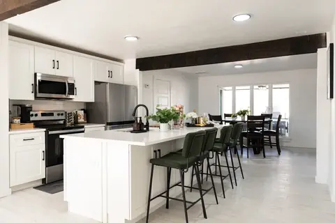 White kitchen with stainless appliances and a long quartz island lined with four green bar stools, opening to the dining area at Will Rogers Manor, Oklahoma City