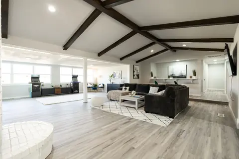 Vaulted great room with dark wood beams, charcoal sectional on a white geometric rug, and a game area with two arcade cabinets beyond at Will Rogers Manor, Oklahoma City